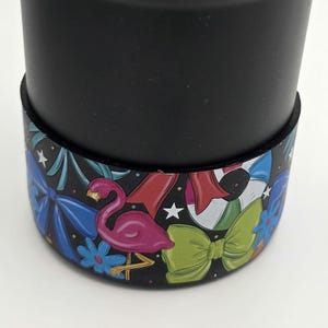 May include: Black tumbler with a colorful band featuring a pink flamingo, blue bows, green bow, and a beach ball design. The band also includes white stars and blue flowers. The tumbler is on a white surface.