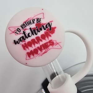 Peut inclure: Un bouchon de paille blanc et rose avec le texte "I'd rather be watching HORROR MOVIES" en noir et rouge. Le bouchon a une boucle pour un retrait facile et est fixé à une paille transparente.