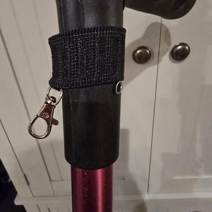 May include: A black fabric strap attached to a dark red pole, featuring a silver metal lobster clasp. The strap is secured around the upper section of the pole, which has a black handle at the top and a small silver rivet visible on the side.