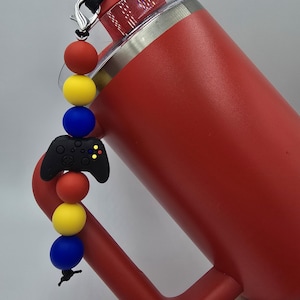 Könnte beinhalten: Ein roter isolierter Becher mit Griff und durchsichtigem Deckel. Ein Perlenanhänger mit einem schwarzen Videospiel-Controller und roten, gelben und blauen Perlen ist am Becher befestigt.