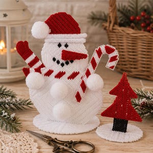 May include: A white plastic canvas snowman figure wearing a red hat and scarf, holding a red and white striped candy cane, accented with white pom-poms. Beside it stands a small red Christmas tree on a white base, with metal scissors nearby.