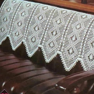 May include: A white, crocheted sofa cover with a geometric diamond pattern. The cover has a scalloped edge and is draped over a dark brown leather sofa. The design features repeating diamond shapes.