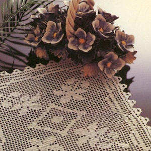 May include: A cream-colored, crocheted doily with a scalloped edge and a floral pattern. A basket of purple flowers with a woven handle sits atop the doily. Green palm fronds are visible in the background.