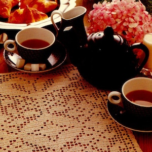 May include: A crocheted table runner with a delicate floral pattern in a beige color. The runner is placed on a wooden table with a teapot, cups, and pastries.