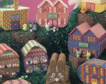 May include: A collection of colorful plastic canvas Easter egg village designs. The designs include houses, a basket, and a bunny, all crafted with needlework. The text on the cover reads "Easter Egg Village" and "plastic canvas".