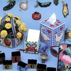 May include: A variety of plastic canvas crafts are displayed, such as a flower basket, tissue box, and lettered boxes. The image showcases needlepoint designs in the shapes of food and animals, with the text "plastic canvas BAZAAR" at the top.