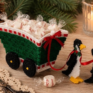 May include: A green and white plastic canvas wagon filled with individually wrapped white candies, pulled by three crocheted penguins in black, white, and yellow. The penguins are connected by a red ribbon with small green holly accents.