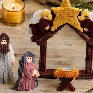 Puede incluir: Un set de nacimiento de lienzo plástico con un marco de establo marrón coronado por una estrella amarilla, dos ángeles con alas blancas, una figura con túnica gris, una con túnica rosa y un pequeño pesebre naranja. La estrella contiene texto.