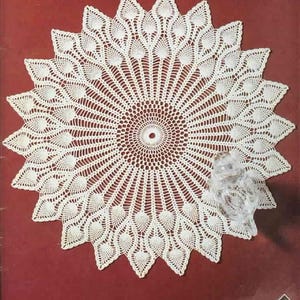 May include: A white, circular doily with a detailed, openwork design. The doily features a central circle with radiating lines and a scalloped edge. The design includes leaf-like patterns. A clear glass owl figurine is on the right side.
