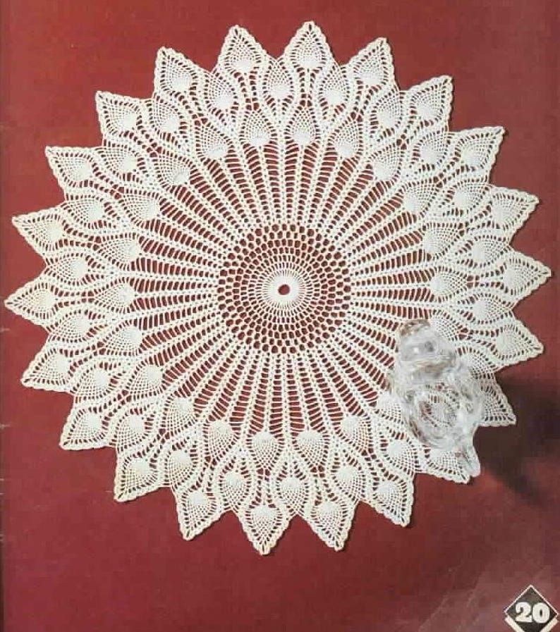 May include: A white, circular doily with a detailed, openwork design. The doily features a central circle with radiating lines and a scalloped edge. The design includes leaf-like patterns. A clear glass owl figurine is on the right side.