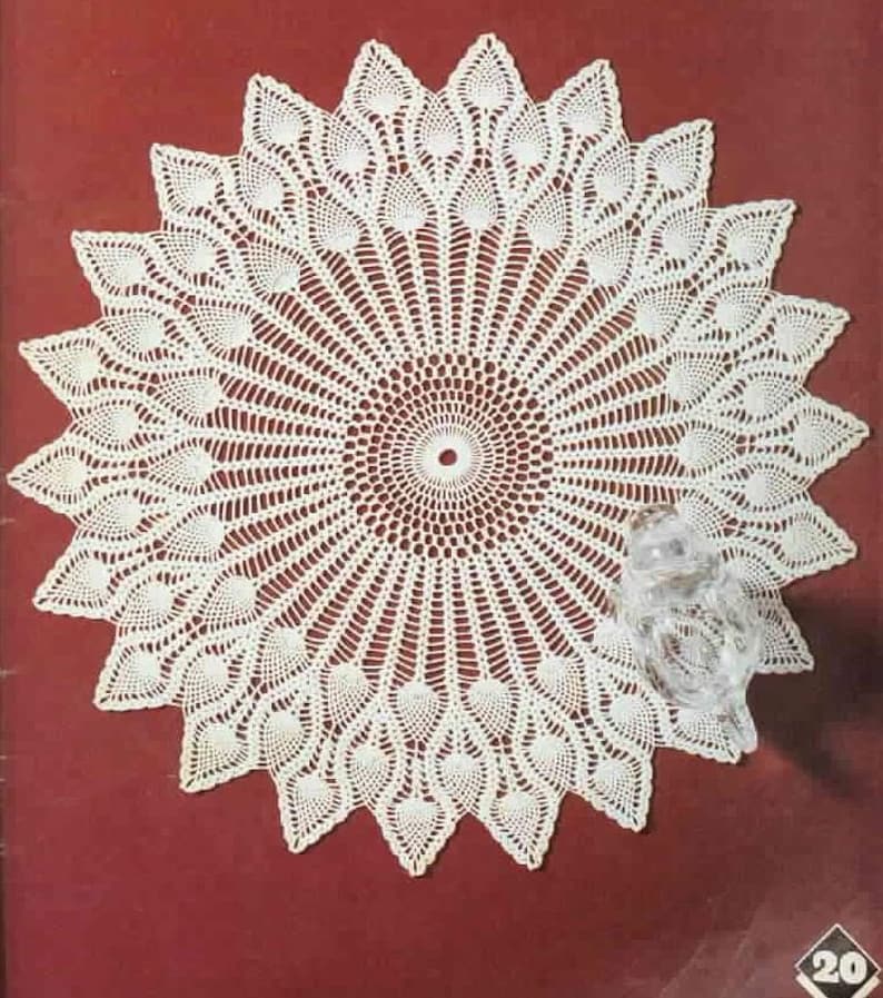 May include: A white, circular doily with a detailed, openwork design. The doily features a central circle with radiating lines and a scalloped edge. The design includes leaf-like patterns. A clear glass owl figurine is on the right side.