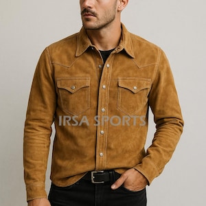 May include: A tan suede shirt with two chest pockets and snap closures. The shirt has a classic collar and long sleeves. The text "IRSA SPORTS" is printed on the front. The person is wearing black jeans and a black belt.