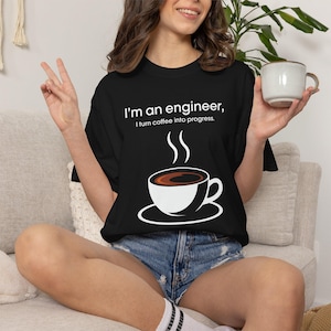 May include: Black t-shirt with the text "I'm an engineer, I turn coffee into progress." A white graphic of a coffee cup with steam rising is below the text. The person is wearing denim shorts and holding a white coffee mug.