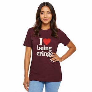 May include: Burgundy crew neck t-shirt with the text "I ♥ being cringe" in white and a red heart. The shirt is paired with blue jeans. The model is smiling and has one hand on their hip.