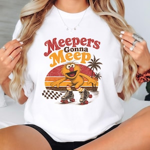 May include: A white t-shirt featuring a graphic print of a fuzzy orange creature on roller skates. The text reads Meepers Gonna Meep above the creature, which is set against a sunset background with palm trees and a checkered pattern at the bottom.