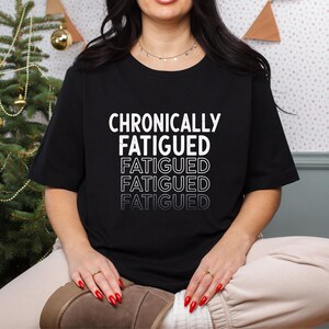 Könnte beinhalten: Ein schwarzes Kurzarm-T-Shirt mit dem weißen Text CHRONICALLY FATIGUED oben, gefolgt von dem Wort FATIGUED, das dreimal darunter mit abnehmender Schriftstärke und Deckkraft wiederholt wird, was einen Verblassungseffekt auf der Brust erzeugt.