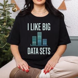 Può includere: Una t-shirt nera a maniche corte con la scritta I LIKE BIG DATA SETS in lettere azzurre. Sotto il testo è presente un grafico a barre con tre colonne di altezze variabili, rese in tonalità di blu e verde acqua sul tessuto nero.
