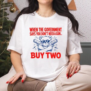 May include: A white short-sleeved t-shirt featuring a graphic of a bear wearing sunglasses and holding two handguns. The red text reads: WHEN THE GOVERNMENT SAYS YOU DON'T NEED A GUN... BUY TWO. The bear and guns are rendered in blue ink.