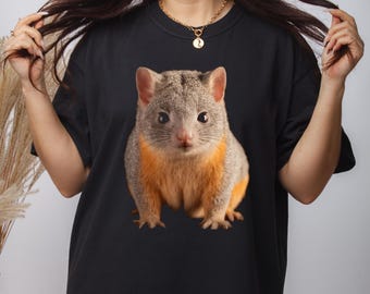 Pode incluir: Uma camiseta preta com uma estampa gráfica de um pequeno animal peludo com pelo laranja no peito e na barriga. O animal tem olhos grandes e escuros e um nariz pontudo.