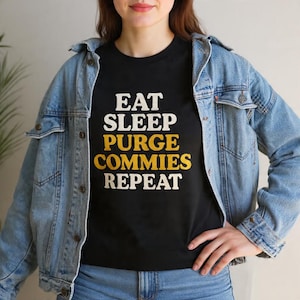 Może przedstawiać: Czarny t-shirt z napisem "EAT SLEEP PURGE COMMIES REPEAT" w białych i żółtych literach. Koszulka jest noszona z jasnoniebieską kurtką dżinsową.