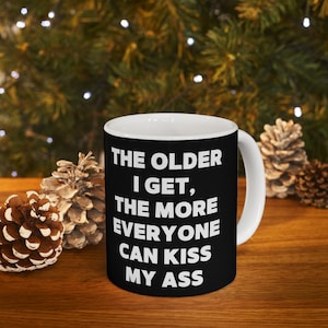 May include: A white ceramic mug with a black exterior featuring the text: "THE OLDER I GET, THE MORE EVERYONE CAN KISS MY ASS" in white. The mug has a white handle and is set against a background of pinecones and greenery.