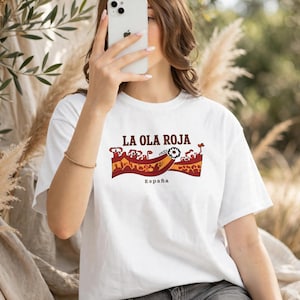 Può includere: Una t-shirt bianca a maniche corte con una stampa grafica che riporta il testo LA OLA ROJA ed España. Il design include un motivo a onda stilizzato rosso e arancione con piccole illustrazioni di persone e un fiore sopra l'onda.