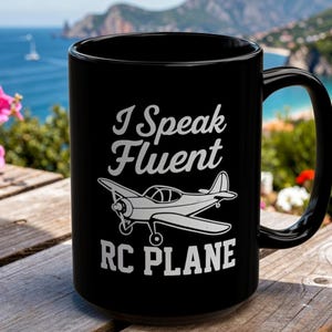 Könnte beinhalten: Ein schwarzer Keramikbecher mit weißem Text, der I Speak Fluent RC PLANE lautet, begleitet von einer weißen grafischen Abbildung eines kleinen Propellerflugzeugs, das mittig auf der Vorderseite des Bechers platziert ist, mit einem geschwungenen Henkel.