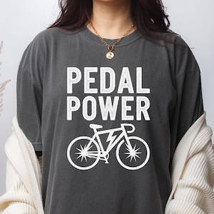 Puede incluir: Camiseta gris oscuro con las palabras blancas "PEDAL POWER" sobre un gráfico de bicicleta blanca con un rayo. La camiseta tiene un corte holgado y está hecha de un material suave.