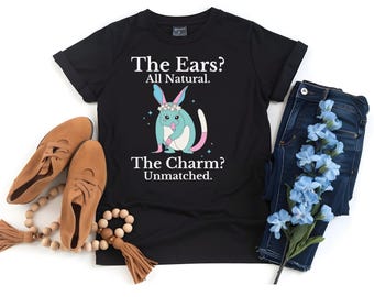May include: Black t-shirt with a graphic of a blue and pink creature with large ears and a flower crown. The text on the shirt reads "The Ears? All Natural. The Charm? Unmatched."