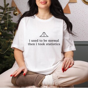 Könnte beinhalten: Ein weißes kurzärmeliges T-Shirt mit einer schwarzen Grafik einer Glockenkurve und dem Text "i used to be normal then i took statistics", der in einer einfachen, klaren Schriftart mittig auf der Brust des Kleidungsstücks gedruckt ist.