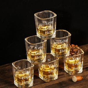 Puede incluir: Seis vasos de chupito cuadrados llenos de líquido ámbar están apilados sobre una superficie de madera. Cada vaso tiene el texto "Your Logo Here" impreso. El fondo es oscuro, con algunos elementos decorativos.