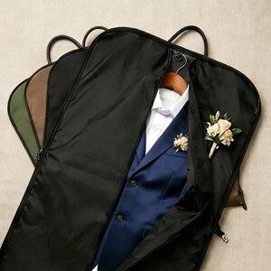 May include: A black garment bag containing a navy blue suit jacket with a white collared shirt and a floral boutonniere pinned to the lapel. Additional garment bags in olive green and brown are layered behind the primary black bag.