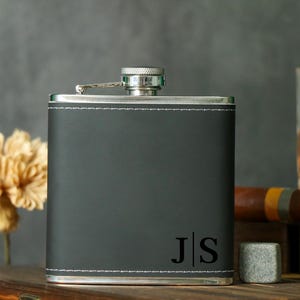 Könnte beinhalten: Eine schwarze, lederbezogene Flasche mit einem silbernen Deckel und einer Monogramm-Inschrift "J|S".