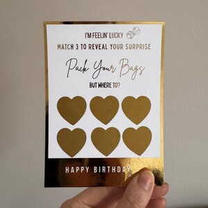 Puede incluir: Una tarjeta de rascar dorada y blanca de cumpleaños con el texto "I'M FEELIN' LUCKY" y "Pack Your Bags". Seis áreas doradas en forma de corazón para rascar son visibles. La tarjeta dice "HAPPY BIRTHDAY".