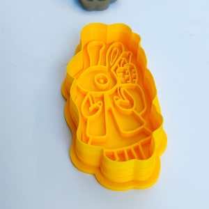 Op de afbeelding: Een gele plastic koekjesvorm in de vorm van een konijn met een mand. Een grijze koekje gemaakt met de vorm is erboven. De vorm staat op een wit oppervlak.