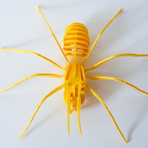 Può includere: Una scultura di insetto giallo brillante con lunghe zampe sottili e un corpo segmentato. L'insetto è composto da pezzi ad incastro, creando un effetto tridimensionale. La scultura è posta su uno sfondo bianco.