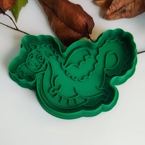 Pode incluir: Um cortador de biscoitos de plástico verde em forma de dragão. O cortador apresenta um design detalhado de um dragão amigável com asas e uma cauda enrolada. O cortador é projetado para fazer biscoitos ou outras guloseimas.