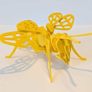 Pode incluir: Um quebra-cabeça de inseto de madeira tridimensional amarelo brilhante. O inseto tem asas, pernas e antenas detalhadas. O quebra-cabeça é montado e projeta uma sombra na superfície branca.