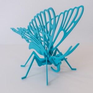 Pode incluir: Uma escultura de borboleta turquesa com intrincados recortes nas asas. A borboleta tem um corpo e pernas detalhados, em pé sobre uma superfície branca. As asas estão abertas, mostrando o design delicado. Adequado para decoração de casa.