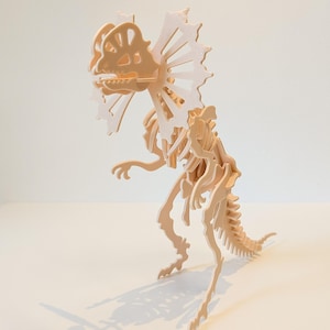Pode incluir: Um quebra-cabeça de esqueleto de dinossauro Dilophosaurus em madeira clara. O quebra-cabeça apresenta um design detalhado dos ossos, da gola e da estrutura corporal do dinossauro. O dinossauro está em uma superfície branca.