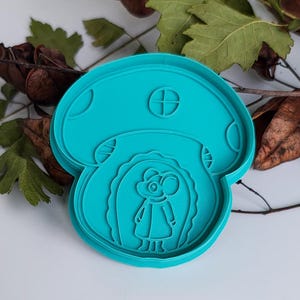 Può includere: Tagliapasta a forma di fungo di colore turchese. Il tagliapasta presenta un topo con un vestito all'interno del fungo. Il fungo ha una piccola finestra con un disegno a croce sulla parte superiore.