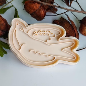 May include: A beige plastic cookie cutter in the shape of a dove with an olive branch. The dove is in a layered design, with the outline of the bird and the branch clearly defined. The cutter is surrounded by dried leaves and branches.