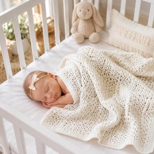 Puede incluir: Una manta de bebé de ganchillo calado en color crema con un patrón geométrico texturizado. La manta cubre a un bebé dormido que lleva una diadema fina de color crema con un pequeño lazo. Un juguete de peluche de conejo marrón claro está cerca.