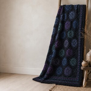 May include: A crochet blanket featuring a repeating pattern of hexagonal motifs in shades of deep blue, purple, and teal, framed by a black border. The blanket has a textured surface with intricate floral-like designs and a scalloped edge finish.