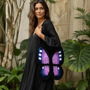 May include: A crochet shoulder bag shaped like a butterfly with purple and lavender wings, accented by dark blue borders and small white spots. The bag features a black crocheted strap and is worn over a plain black dress with a V-neckline.