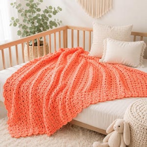 Peut inclure: Une couverture au crochet de couleur corail vif avec un motif texturé à mailles ouvertes et un bord festonné. Le jeté rectangulaire est drapé sur un drap de lit blanc, présentant un motif vertical répétitif de boucles et de petits espaces.