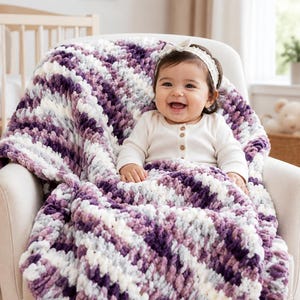 Könnte beinhalten: Eine grob gestrickte Babydecke mit einem melierten Muster aus dunkelviolettem, helllavendelfarbenem, grauem und weißem Garn. Die Decke hat eine weiche, strukturierte Optik mit dicken, flauschigen Maschen, die in diagonalen Streifen verlaufen.