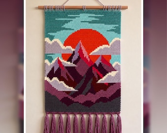 May include: A rectangular crochet wall hanging featuring a mountain landscape with a large orange sun, purple and dark gray peaks, and light blue sky. The piece is suspended from a wooden dowel with a string and finished with purple tassels.