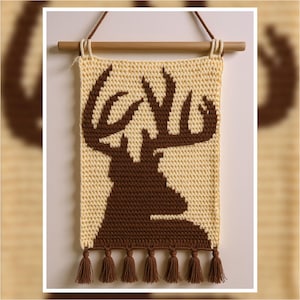 May include: A handmade wall hanging featuring a crocheted design of a deer silhouette in brown against a cream background. The piece is suspended from a wooden dowel with brown tassels at the bottom, creating a rustic, decorative element.
