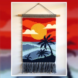 Pode incluir: Uma tapeçaria de parede tecida que retrata uma cena de pôr do sol com um grande sol amarelo, nuvens brancas e palmeiras. A obra de arte apresenta um gradiente de cores, incluindo vermelho, laranja, azul e cinza, com uma franja na parte inferior. A tapeçaria é fixada a um pino de madeira.